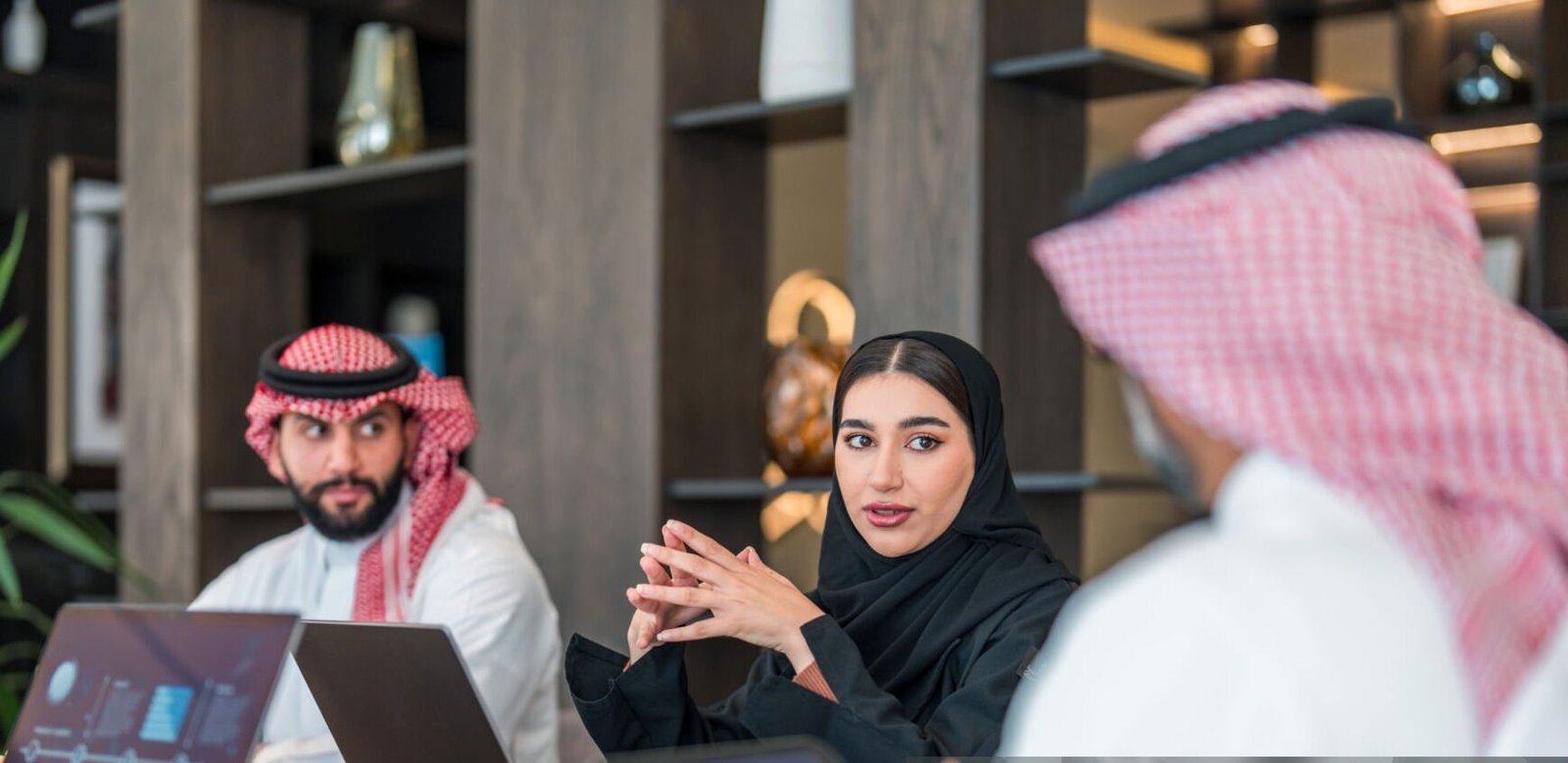 istockphoto-2190756966-2048×2048 Diverse Arabic business executives discuss strategies at a meeting. The man wears traditional attire, while the woman is in a black abaya. Laptops and documents are present in a modern office setting, highlighting professional collaboration and analysis.